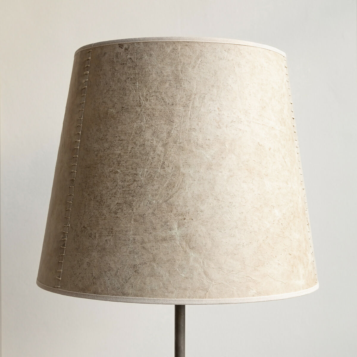 A beige hand stitched tapered lampshade made of Lokta paper with a natural linen trim, displayed on a lamp stand against a white background,  available  to buy on the-salon.com. SALON curates the finest , unique, handmade pieces from the interior industry's best makers and brands. Shop luxury furniture, lighting, rugs, art, antiques and vintage pieces and accessories from SALON interior designed rooms.