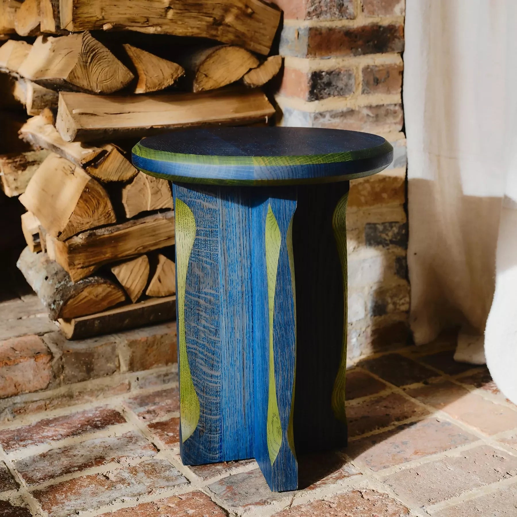 A round, blue with green oak side table with a cross base, placed on a woven mat,  available  to buy on the-salon.com. SALON curates the finest , unique, handmade pieces from the interior industry's best makers and brands. Shop luxury furniture, lighting, rugs, art, antiques and vintage pieces and accessories from SALON interior designed rooms.