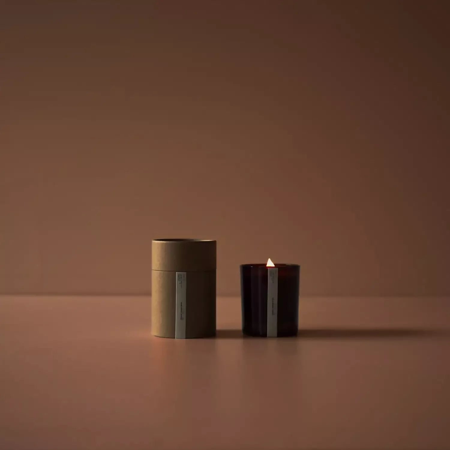A candle with a black holder, featuring a lit flame, placed next to its cylindrical packaging on a plain background, available  to buy on the-salon.com. SALON curates the finest , unique, handmade pieces from the interior industry's best makers and brands. Shop luxury furniture, lighting, rugs, art, antiques and vintage pieces and accessories from SALON interior designed rooms.