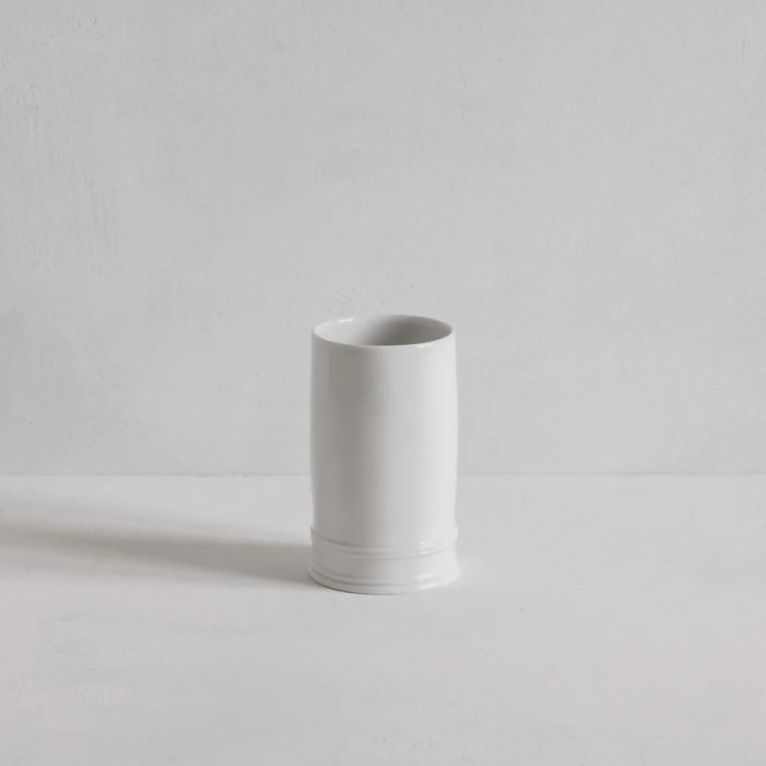 A white, cylindrical, porcelain column vase with an unglazed exterior, displayed against a neutral background,  available  to buy on the-salon.com. SALON curates the finest , unique, handmade pieces from the interior industry's best makers and brands. Shop luxury furniture, lighting, rugs, art, antiques and vintage pieces and accessories from SALON interior designed rooms.