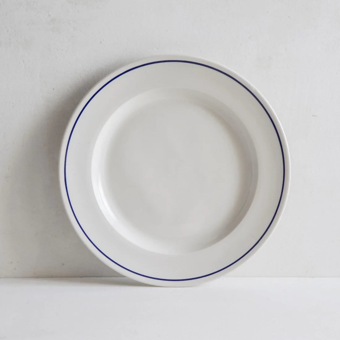 A white porcelain dinner plate with a rich cobalt blue line along the rim, placed against a neutral background, available  to buy on the-salon.com. SALON curates the finest , unique, handmade pieces from the interior industry's best makers and brands. Shop luxury furniture, lighting, rugs, art, antiques and vintage pieces and accessories from SALON interior designed rooms.