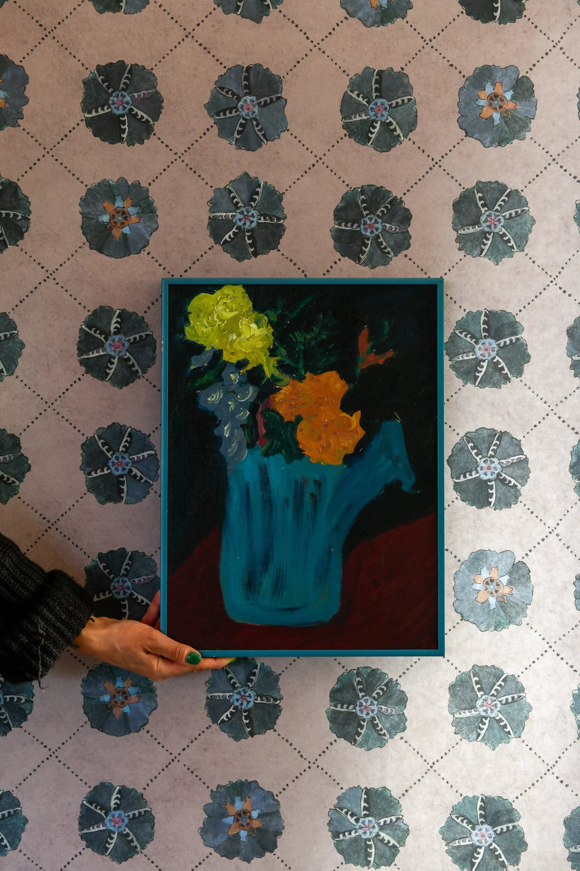 A person holding a swatch of SUZANI wallpaper with a geometric pattern featuring a blue vase with yellow flowers in the center available  to buy on the-salon.com. SALON curates the finest , unique, handmade pieces from the interior industry's best makers and brands. Shop luxury furniture, lighting, rugs, art, antiques and vintage pieces and accessories from SALON interior designed rooms.