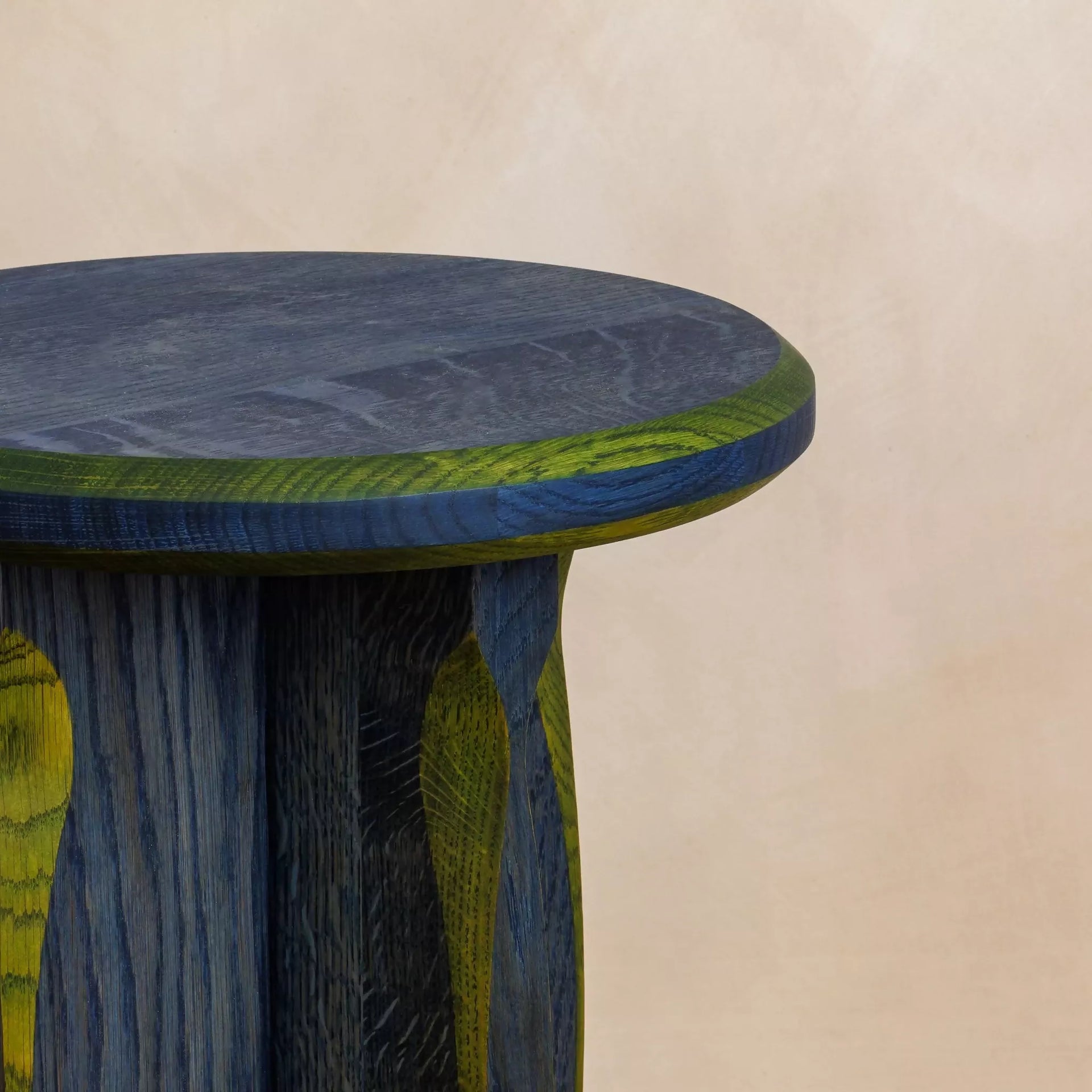 A round, blue with green oak side table with a cross base, placed on a woven mat,  available  to buy on the-salon.com. SALON curates the finest , unique, handmade pieces from the interior industry's best makers and brands. Shop luxury furniture, lighting, rugs, art, antiques and vintage pieces and accessories from SALON interior designed rooms.