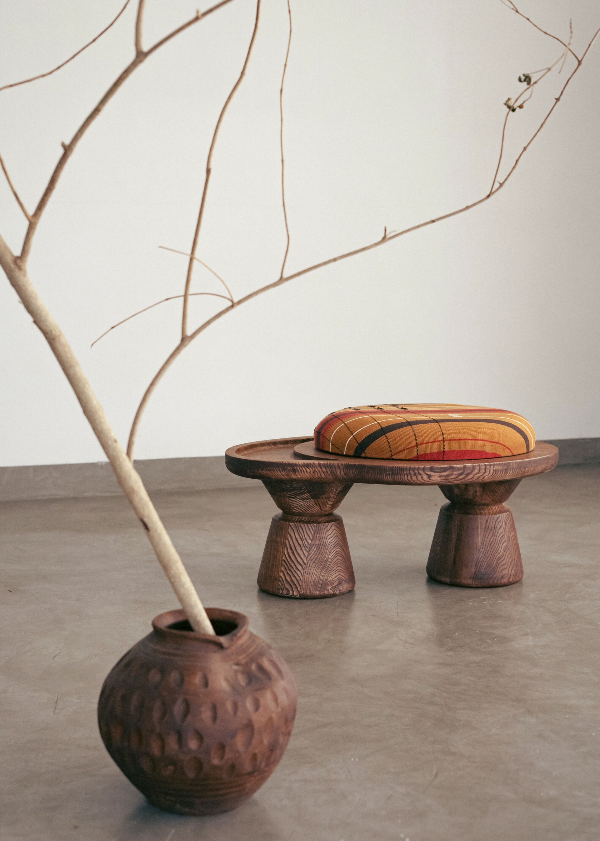 A sculptural single-seater ottoman with a curved body, upholstered in mustard color, placed next to a large ceramic pot with a branch in it, on a concrete floor,  available  to buy on the-salon.com. SALON curates the finest , unique, handmade pieces from the interior industry's best makers and brands. Shop luxury furniture, lighting, rugs, art, antiques and vintage pieces and accessories from SALON interior designed rooms.