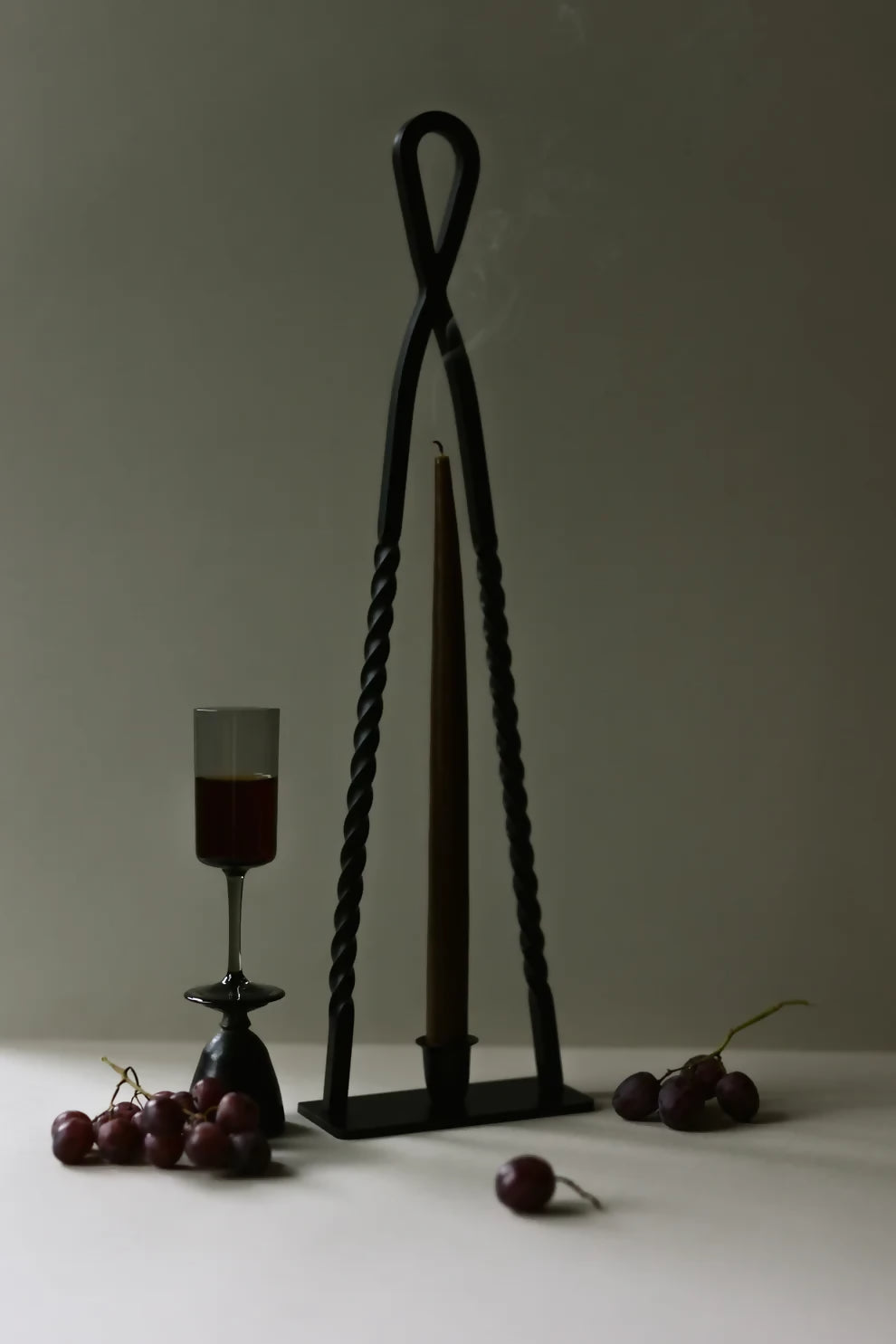 A black, handmade steel Alchemy candle holder by HUEY, displayed on a gray background with a candle inside it, surrounded by cherries on the table, available  to buy on the-salon.com. SALON curates the finest , unique, handmade pieces from the interior industry's best makers and brands. Shop luxury furniture, lighting, rugs, art, antiques and vintage pieces and accessories from SALON interior designed rooms.