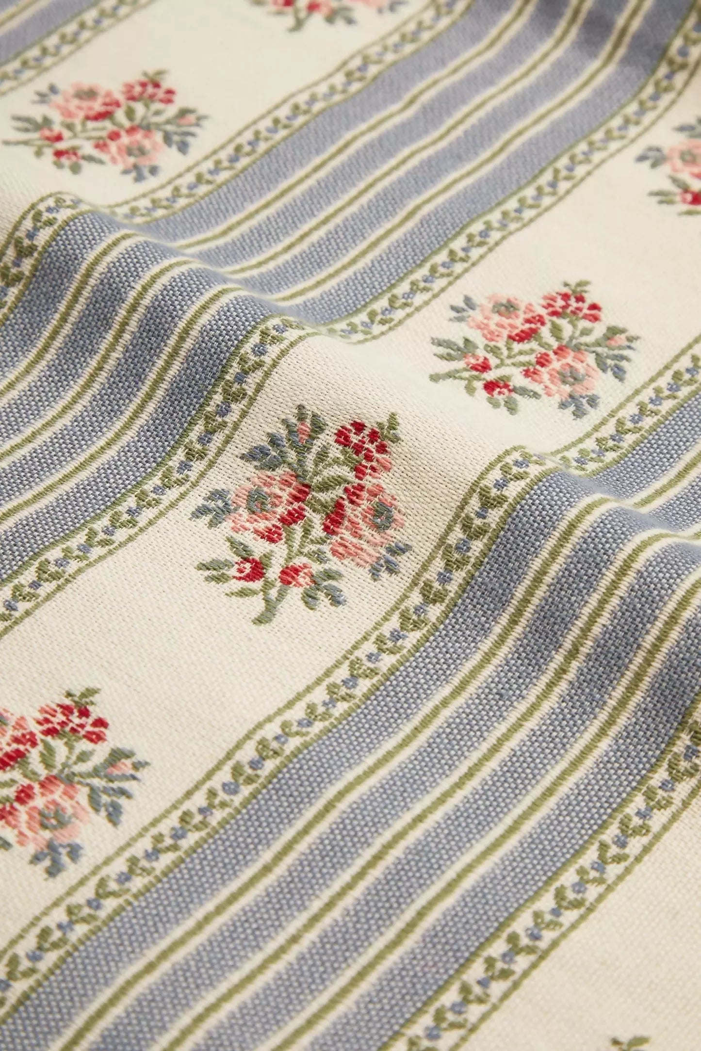 Close-up view of a fabric featuring a jacquard weave with a pattern of delicate embroidered floral motifs, stripes, and leaves in blue, green, and beige colours,  available  to buy on the-salon.com. SALON curates the finest , unique, handmade pieces from the interior industry's best makers and brands. Shop luxury furniture, lighting, rugs, art, antiques and vintage pieces and accessories from SALON interior designed rooms.