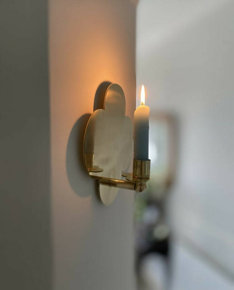 A solid brass wall-mounted candle sconce in the shape of a quatrefoil with a lit candle, reflecting light on a wall,  available  to buy on the-salon.com. SALON curates the finest , unique, handmade pieces from the interior industry's best makers and brands. Shop luxury furniture, lighting, rugs, art, antiques and vintage pieces and accessories from SALON interior designed rooms.