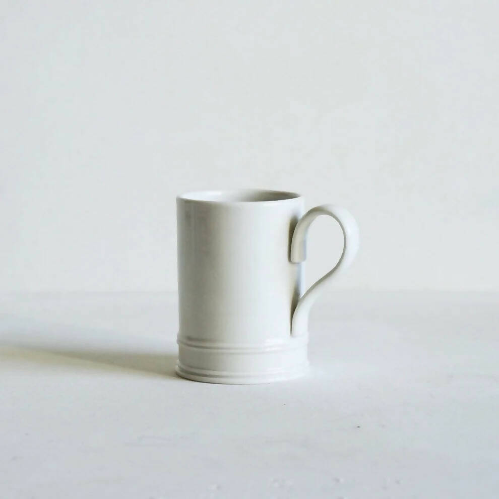 A white porcelain coffee mug with a handle, on a plain background, available  to buy on the-salon.com. SALON curates the finest , unique, handmade pieces from the interior industry's best makers and brands. Shop luxury furniture, lighting, rugs, art, antiques and vintage pieces and accessories from SALON interior designed rooms.