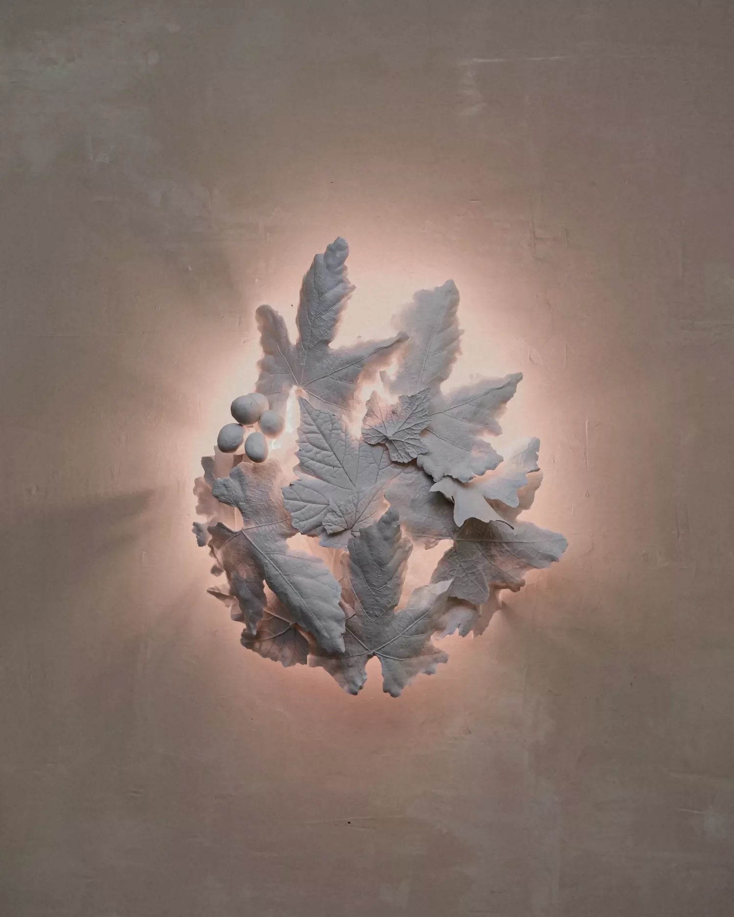A sculptural wall light fixture featuring white leaf and berry motifs mounted on a warm beige plaster wall, casting a soft glow.