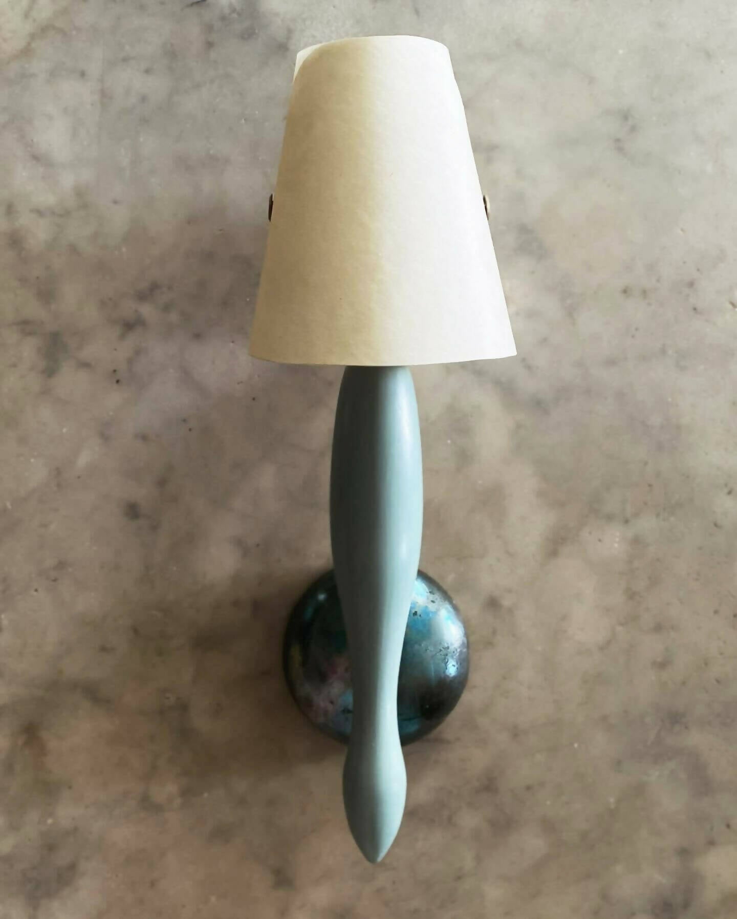 A wall light fixture with a blue, stem-like structure and a white conical shade, mounted on a blue-colored plate, available  to buy on the-salon.com. SALON curates the finest , unique, handmade pieces from the interior industry's best makers and brands. Shop luxury furniture, lighting, rugs, art, antiques and vintage pieces and accessories from SALON interior designed rooms.