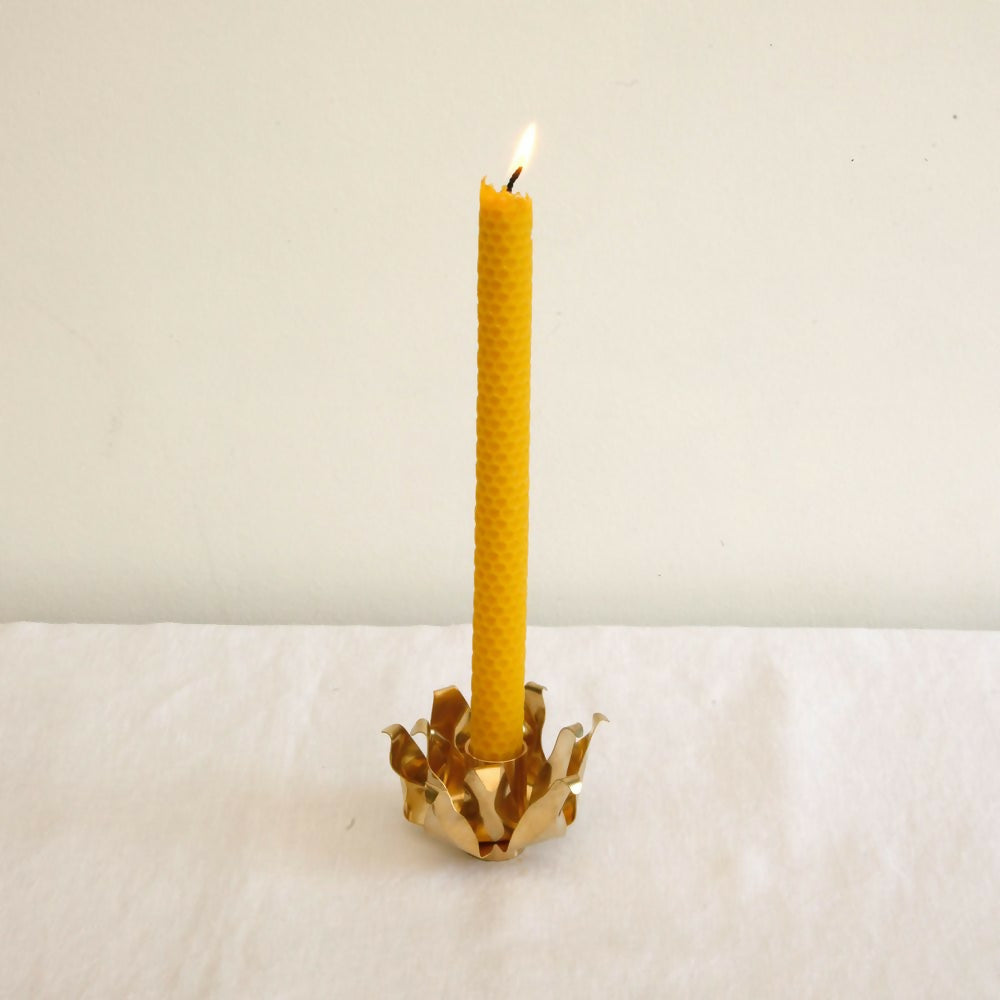 A brass candle holder with a floral design, holding a yellow candle, placed on a white surface,  available  to buy on the-salon.com. SALON curates the finest , unique, handmade pieces from the interior industry's best makers and brands. Shop luxury furniture, lighting, rugs, art, antiques and vintage pieces and accessories from SALON interior designed rooms.