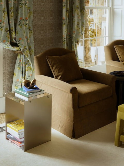 A brown upholstered armchair with classic curved arms and a plump cushion, placed in a room with floral curtains and a side table with books,  available  to buy on the-salon.com. SALON curates the finest , unique, handmade pieces from the interior industry's best makers and brands. Shop luxury furniture, lighting, rugs, art, antiques and vintage pieces and accessories from SALON interior designed rooms.