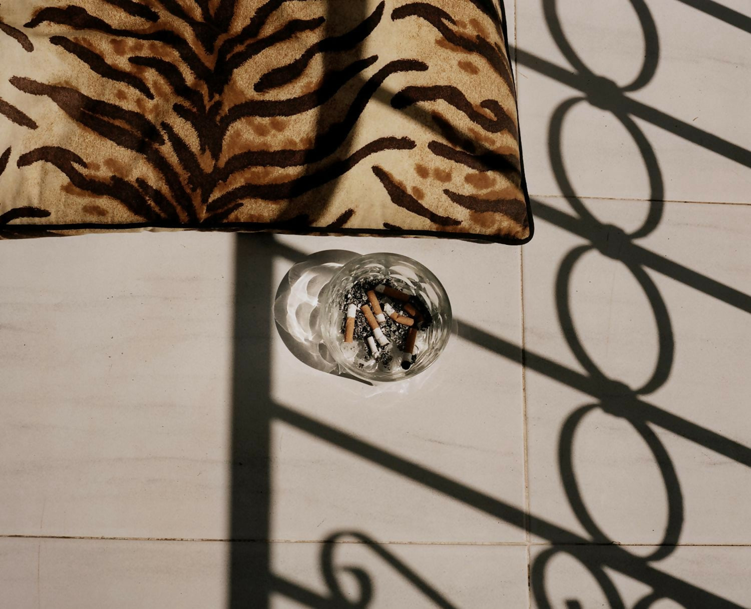 A photograph featuring a close-up of a glass ashtray with cigarettes on a table, surrounded by patterned fabric,  available  to buy on the-salon.com. SALON curates the finest , unique, handmade pieces from the interior industry's best makers and brands. Shop luxury furniture, lighting, rugs, art, antiques and vintage pieces and accessories from SALON interior designed rooms.