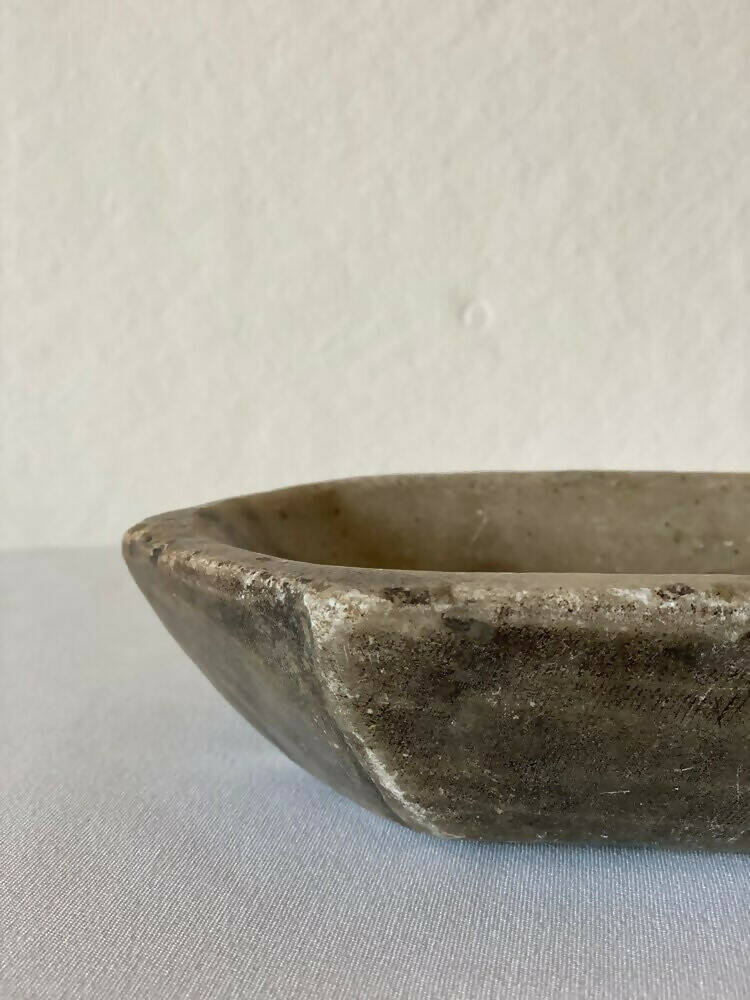 A primitive carved marble bowl with a hexagonal shape, predominantly gray in color, showcasing natural stone patterns, available  to buy on the-salon.com. SALON curates the finest , unique, handmade pieces from the interior industry's best makers and brands. Shop luxury furniture, lighting, rugs, art, antiques and vintage pieces and accessories from SALON interior designed rooms.