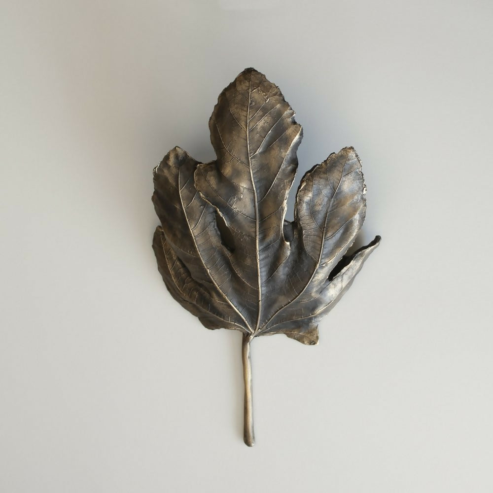 A bronze wall light fixture designed to resemble a fig leaf, with a textured surface and a wall mounting bracket,  available  to buy on the-salon.com. SALON curates the finest , unique, handmade pieces from the interior industry's best makers and brands. Shop luxury furniture, lighting, rugs, art, antiques and vintage pieces and accessories from SALON interior designed rooms.