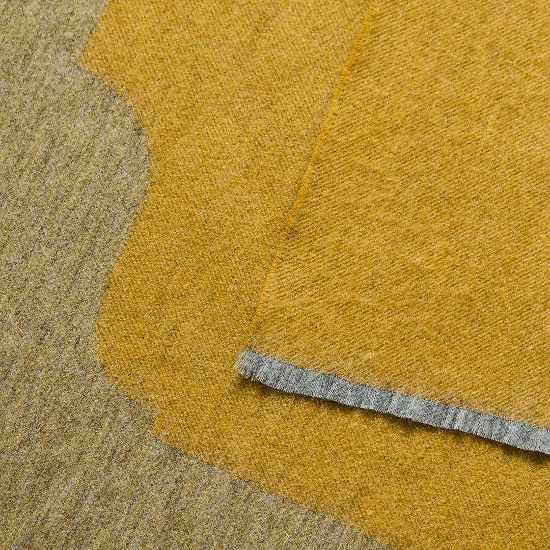 A gold-colored cashmere throw blanket with a decorative border, displaying a small blue and silver brand tag on the bottom right corner,  available  to buy on the-salon.com. SALON curates the finest , unique, handmade pieces from the interior industry's best makers and brands. Shop luxury furniture, lighting, rugs, art, antiques and vintage pieces and accessories from SALON interior designed rooms.