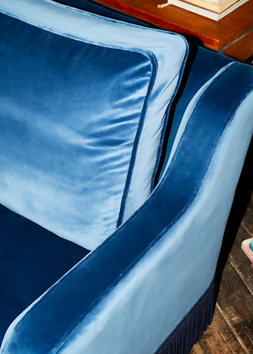 A blue velvet sofa with plush seats and a tasseled fringe, named 'Large Atlanta Sofa by Orior', positioned in a room with a bookshelf, a chair, and a decorative rug,  available  to buy on the-salon.com. SALON curates the finest , unique, handmade pieces from the interior industry's best makers and brands. Shop luxury furniture, lighting, rugs, art, antiques and vintage pieces and accessories from SALON interior designed rooms.