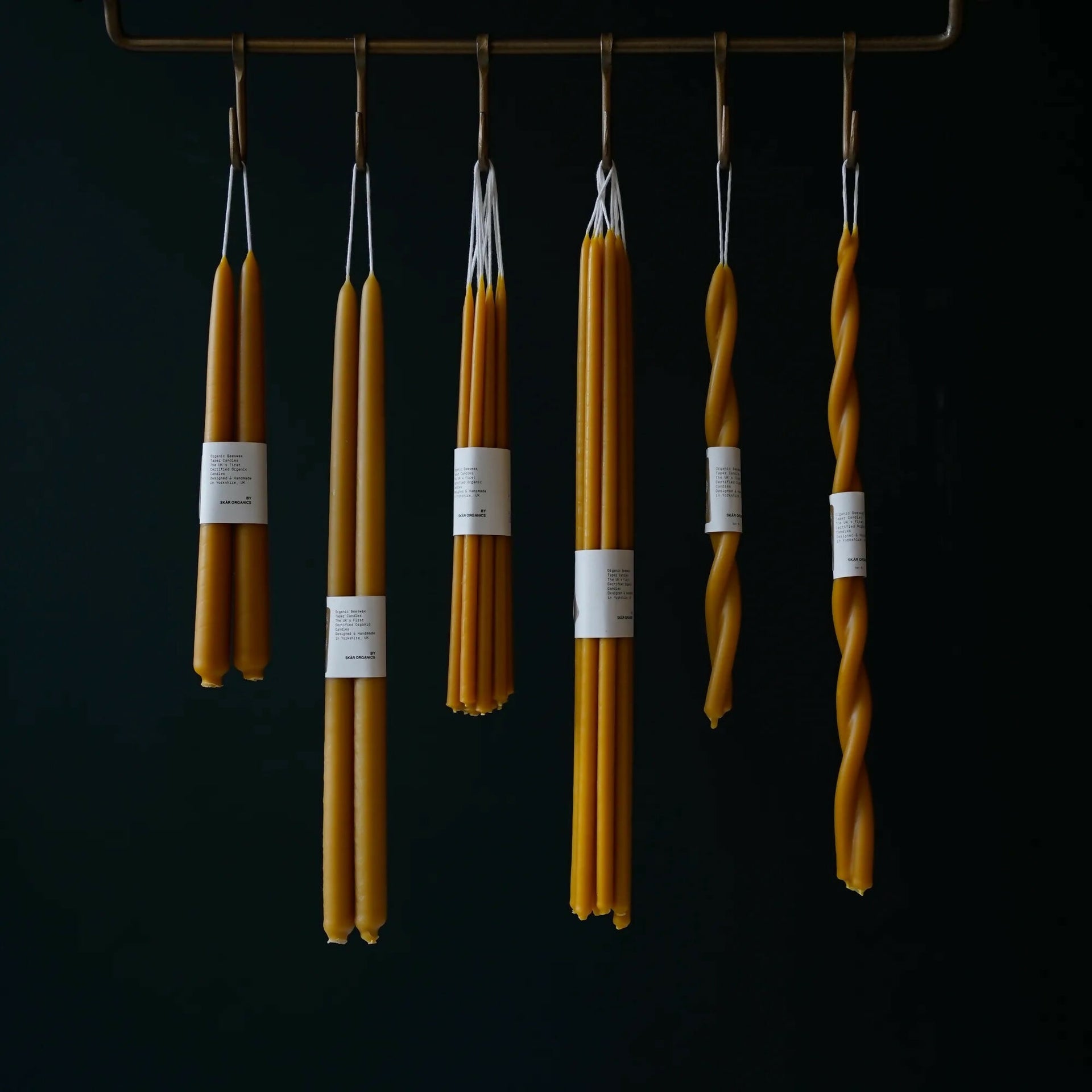 A group of unlit organic beeswax dinner candles, in various shapes including straight and twisted, hanging from a metal rod,  available  to buy on the-salon.com. SALON curates the finest , unique, handmade pieces from the interior industry's best makers and brands. Shop luxury furniture, lighting, rugs, art, antiques and vintage pieces and accessories from SALON interior designed rooms.
