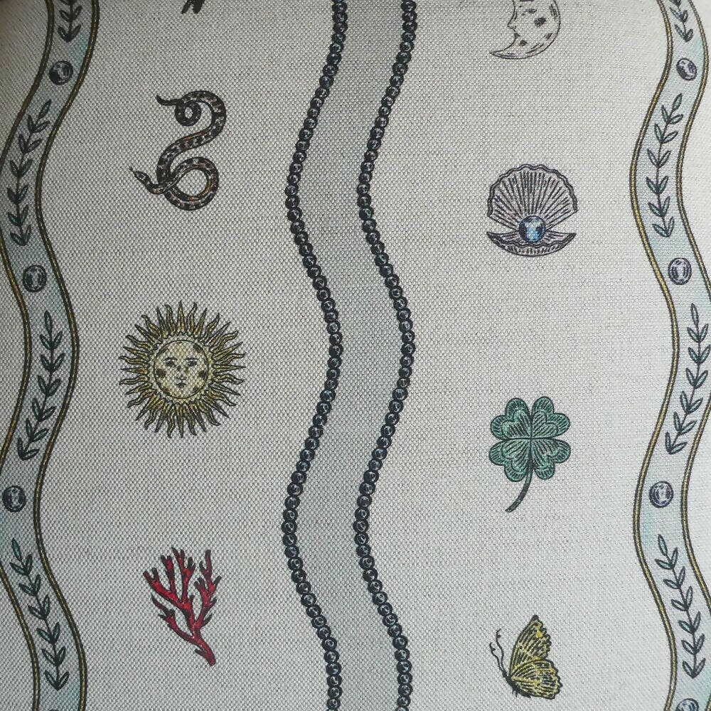 A detailed image of a fabric with a natural and whimsical pattern design, featuring elements such as a snake, a sun, a shell, a clover, and a butterfly, with a moon and leaf motifs,  available  to buy on the-salon.com. SALON curates the finest , unique, handmade pieces from the interior industry's best makers and brands. Shop luxury furniture, lighting, rugs, art, antiques and vintage pieces and accessories from SALON interior designed rooms.