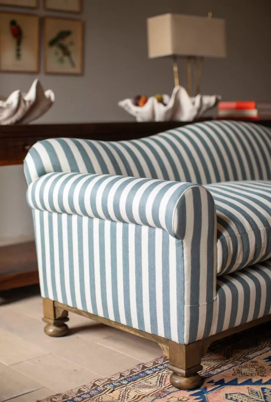 A blue and white striped camel back sofa with solid oak turned legs in a room with decorative items on top, available  to buy on the-salon.com. SALON curates the finest , unique, handmade pieces from the interior industry's best makers and brands. Shop luxury furniture, lighting, rugs, art, antiques and vintage pieces and accessories from SALON interior designed rooms.
