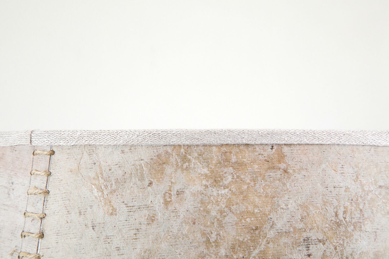 A close up on the stitching on a beige hand stitched tapered lampshade made of Lokta paper with a natural linen trim, against a white background,  available  to buy on the-salon.com. SALON curates the finest , unique, handmade pieces from the interior industry's best makers and brands. Shop luxury furniture, lighting, rugs, art, antiques and vintage pieces and accessories from SALON interior designed rooms.