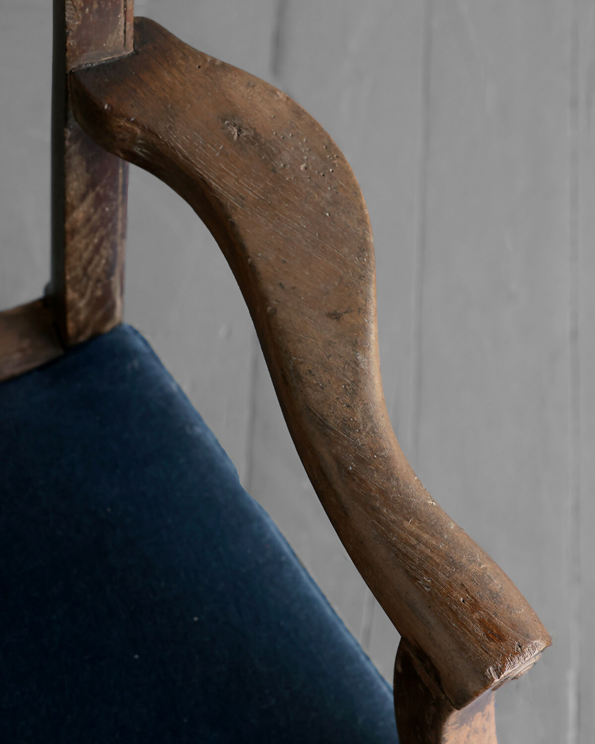 Close-up of a wooden chair armrest with a blue cushion against a gray wall,available to buy from the-salon.com. SALON curates the finest unique handmade pieces from the interior industry's best makers and brands. Shop luxury furniture, lighting, rugs, art, antiques and vintage pieces and accessories from SALON interior designed rooms. 