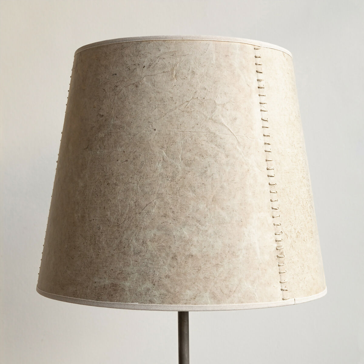 A beige hand stitched tapered lampshade made of Lokta paper with a natural linen trim, displayed on a lamp stand against a white background,  available  to buy on the-salon.com. SALON curates the finest , unique, handmade pieces from the interior industry's best makers and brands. Shop luxury furniture, lighting, rugs, art, antiques and vintage pieces and accessories from SALON interior designed rooms.