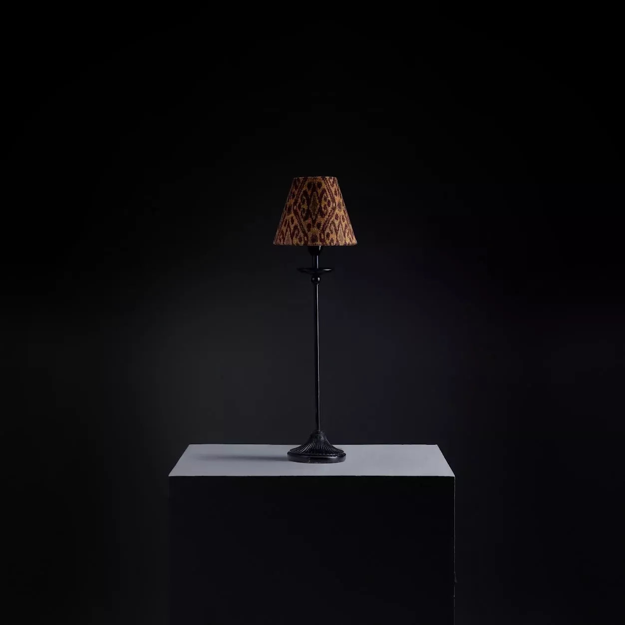 A lampshade with a woven ikat pattern, featuring a geometric design in a tiger's eye color, displayed on a table lamp against a dark background,  available  to buy on the-salon.com. SALON curates the finest , unique, handmade pieces from the interior industry's best makers and brands. Shop luxury furniture, lighting, rugs, art, antiques and vintage pieces and accessories from SALON interior designed rooms.