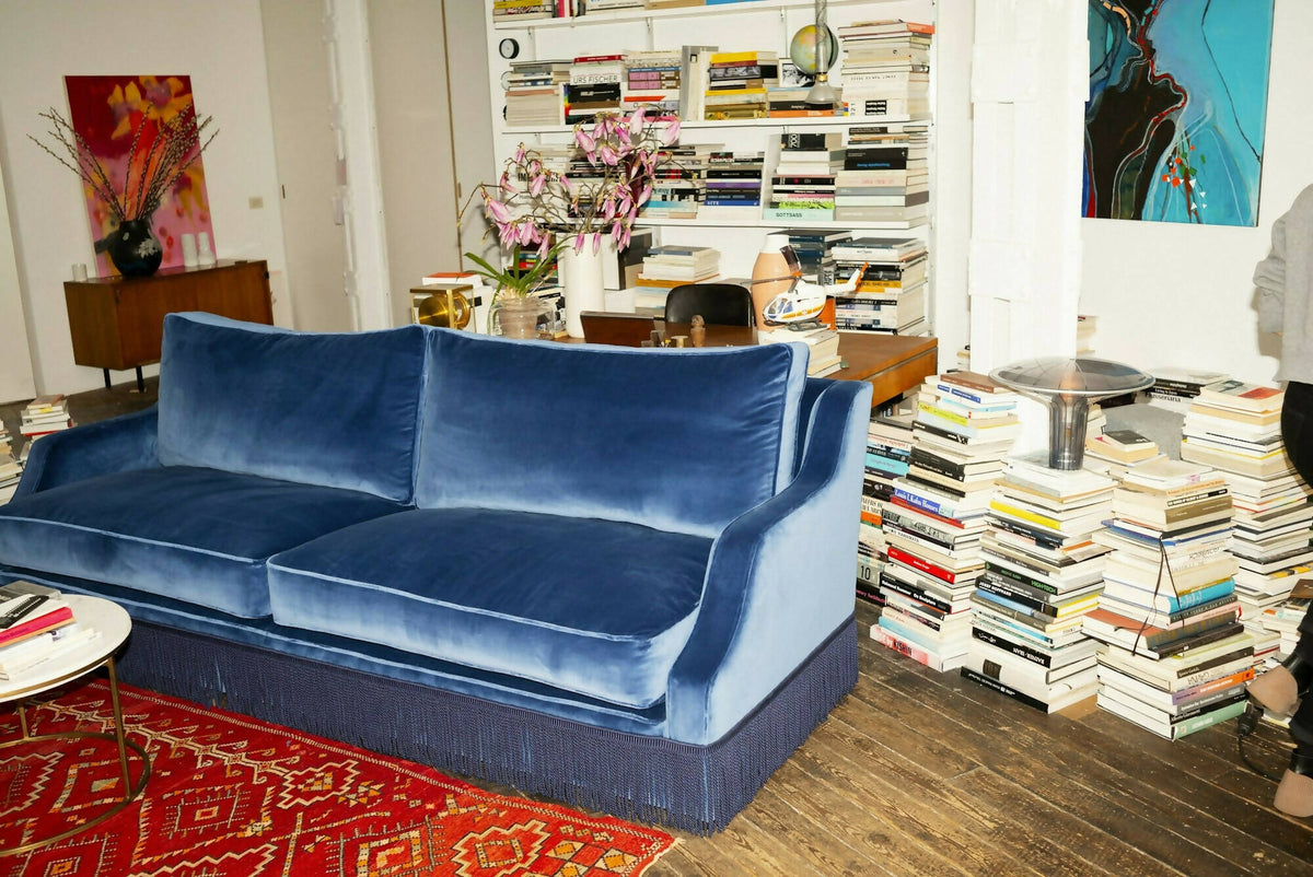 A blue velvet sofa with plush seats and a tasseled fringe, named 'Large Atlanta Sofa by Orior', positioned in a room with a bookshelf, a chair, and a decorative rug,  available  to buy on the-salon.com. SALON curates the finest , unique, handmade pieces from the interior industry's best makers and brands. Shop luxury furniture, lighting, rugs, art, antiques and vintage pieces and accessories from SALON interior designed rooms.