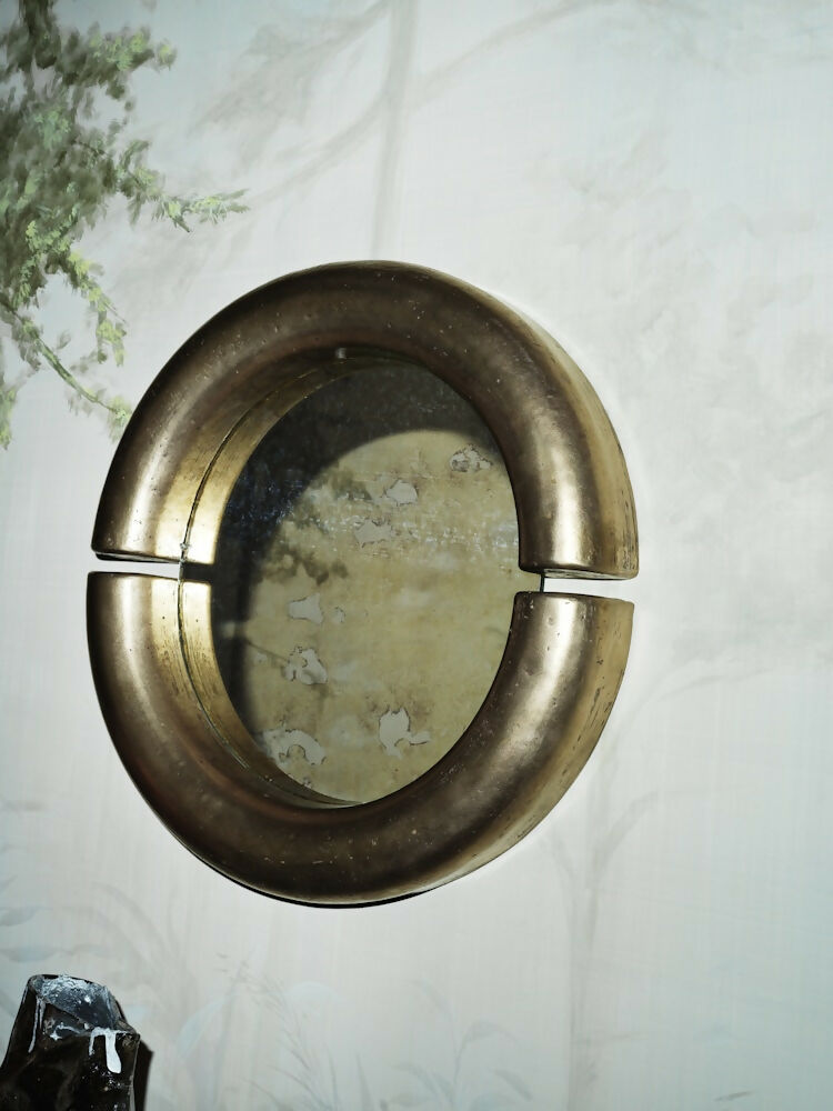 A round wall mirror with a thick, split circular frame in a brushed brass or bronze finish, mounted on a light-colored wall with faint botanical patterns.