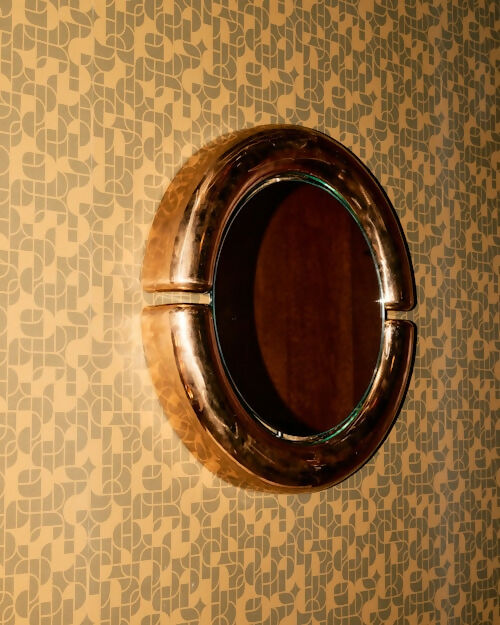 An oval wall mirror with a thick, polished metallic bronze frame mounted on a patterned wallpaper background.