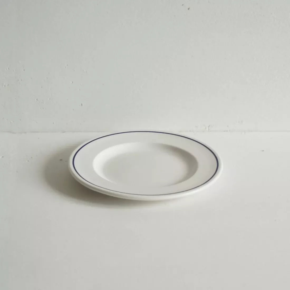 A small round white porcelain side plate with a rich cobalt blue line along the rim, displayed against a white background, available  to buy on the-salon.com. SALON curates the finest , unique, handmade pieces from the interior industry's best makers and brands. Shop luxury furniture, lighting, rugs, art, antiques and vintage pieces and accessories from SALON interior designed rooms.
