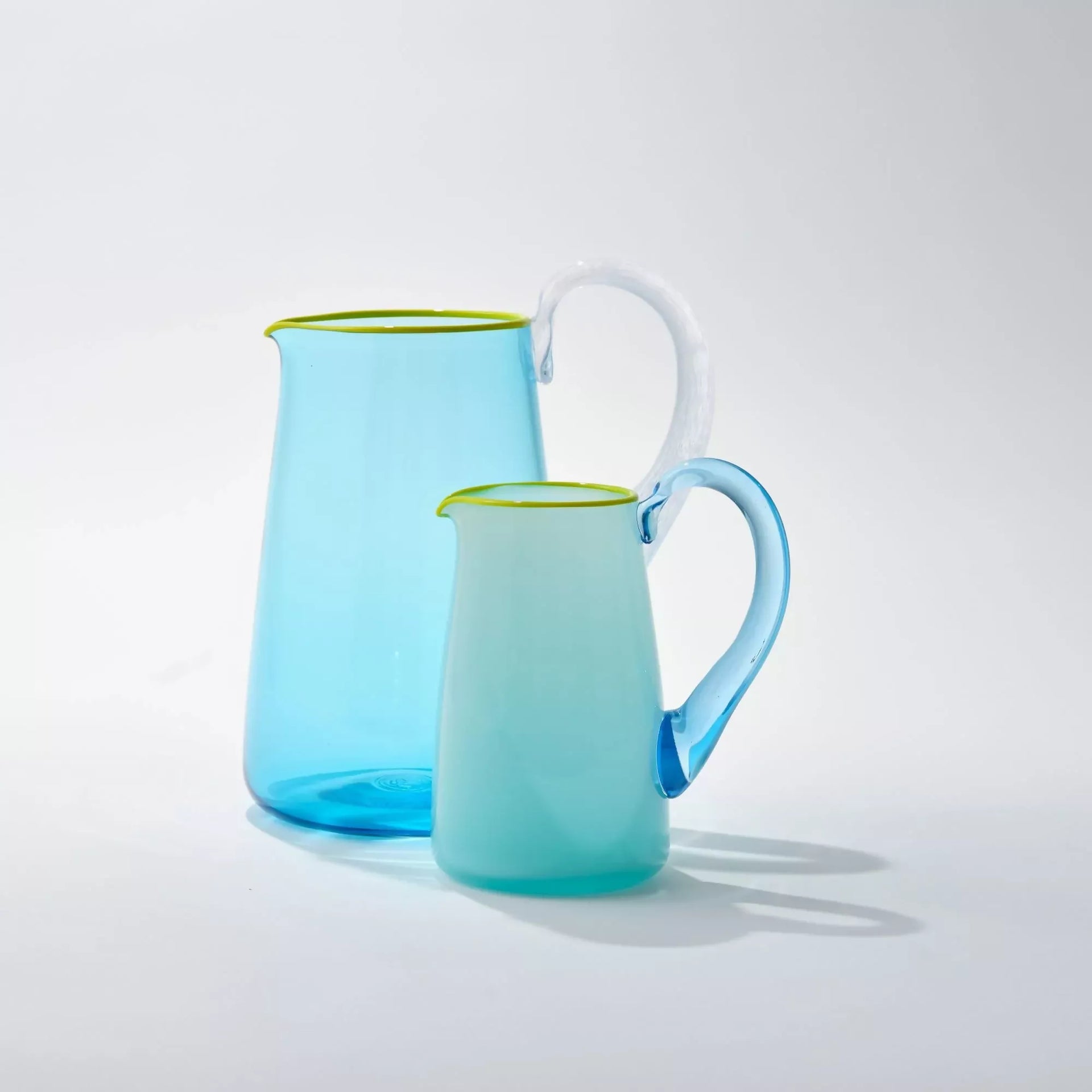 An image of two hand-blown blue glass milk jugs with an aqua blue handle and a celadon lip, against a white background- available  to buy on the-salon.com. SALON curates the finest , unique, handmade pieces from the interior industry's best makers and brands. Shop luxury furniture, lighting, rugs, art, antiques and vintage pieces and accessories from SALON interior designed rooms.