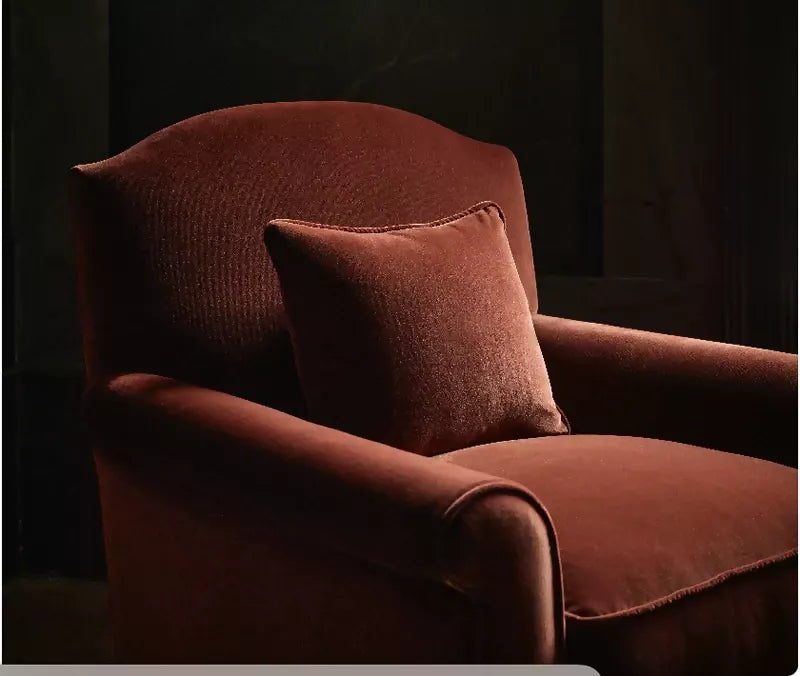 A brown upholstered armchair with classic curved arms and a plump cushion, in the foreground of a dark background with liminal lighting,  available  to buy on the-salon.com. SALON curates the finest , unique, handmade pieces from the interior industry's best makers and brands. Shop luxury furniture, lighting, rugs, art, antiques and vintage pieces and accessories from SALON interior designed rooms.