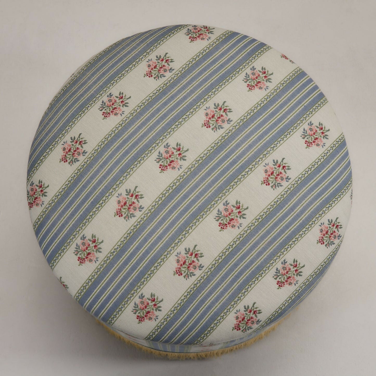 A close up of the  round footstool upholstered in a multicoloured fabric with stripes and floral patterns, featuring a silky champagne fringe at the bottom,  available  to buy on the-salon.com. SALON curates the finest , unique, handmade pieces from the interior industry's best makers and brands. Shop luxury furniture, lighting, rugs, art, antiques and vintage pieces and accessories from SALON interior designed rooms.
