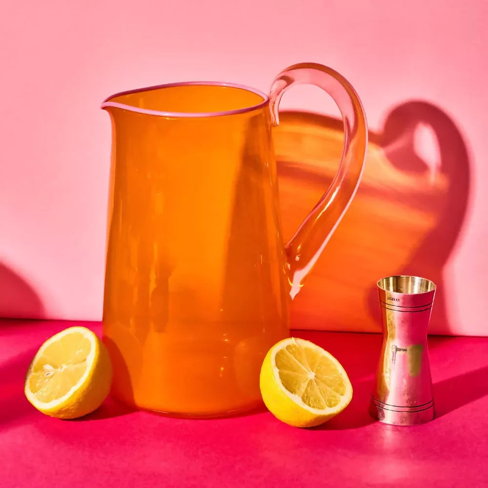 Hand blown, transparent orange glass jug with a blush handle and contrasting pale pink lip on a pink background with two sliced lemons and a shot measuring cup,  available  to buy on the-salon.com. SALON curates the finest , unique, handmade pieces from the interior industry's best makers and brands. Shop luxury furniture, lighting, rugs, art, antiques and vintage pieces and accessories from SALON interior designed rooms.
