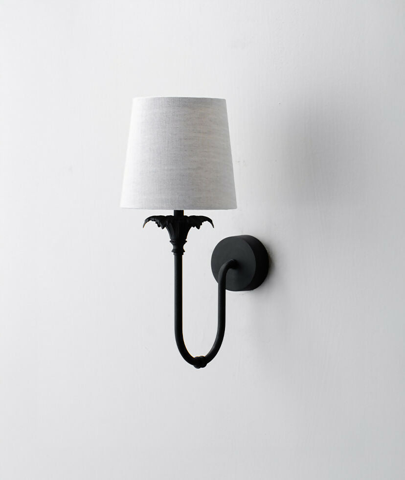 A wall-mounted light fixture with a black-colored metal arm and a white, solid-patterned lampshade,  available  to buy on the-salon.com. SALON curates the finest , unique, handmade pieces from the interior industry's best makers and brands. Shop luxury furniture, lighting, rugs, art, antiques and vintage pieces and accessories from SALON interior designed rooms.