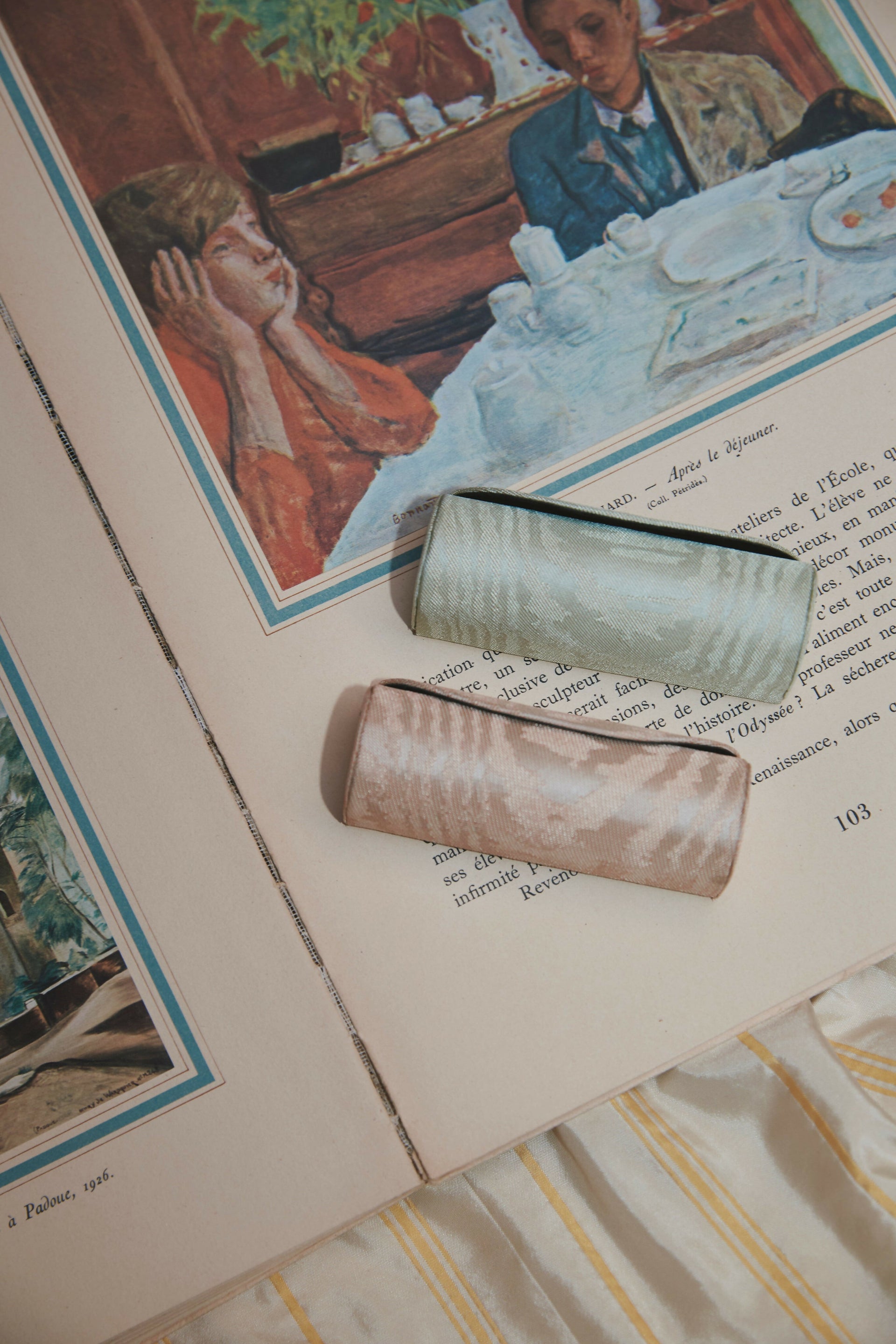 Two cylindrical objects on an open book with a vintage-style illustration of a woman and child in the background,  available  to buy on the-salon.com. SALON curates the finest , unique, handmade pieces from the interior industry's best makers and brands. Shop luxury furniture, lighting, rugs, art, antiques and vintage pieces and accessories from SALON interior designed rooms.