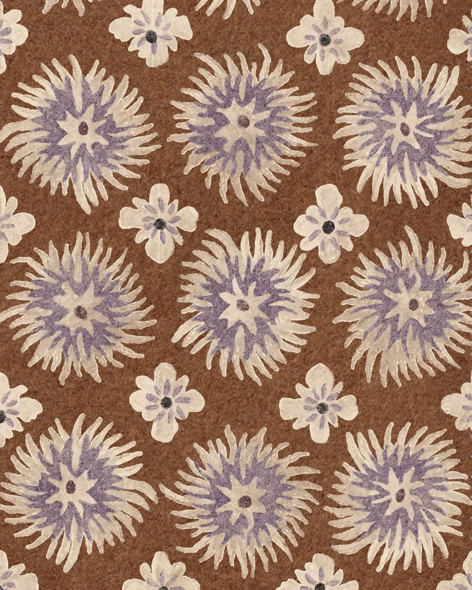 A close-up of a woven fabric or rug swatch with a repeating floral pattern. The design features large circular sunburst motifs in lavender-purple and cream, interspersed with smaller cream flowers, all set against a medium brown background.