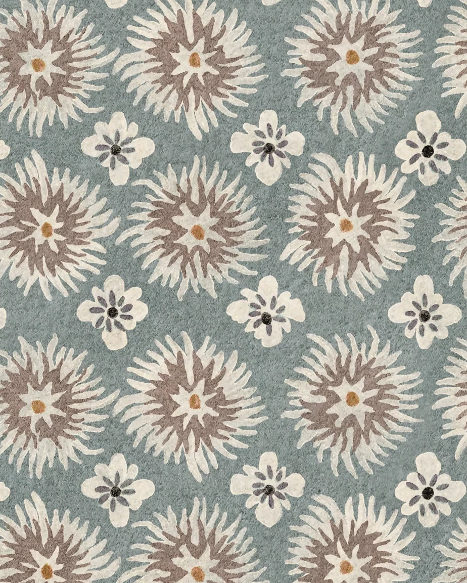 A square fabric or rug swatch with a repeating floral pattern on a muted blue-gray background. The design features large sunburst motifs in taupe and cream, with smaller cream flowers scattered throughout.