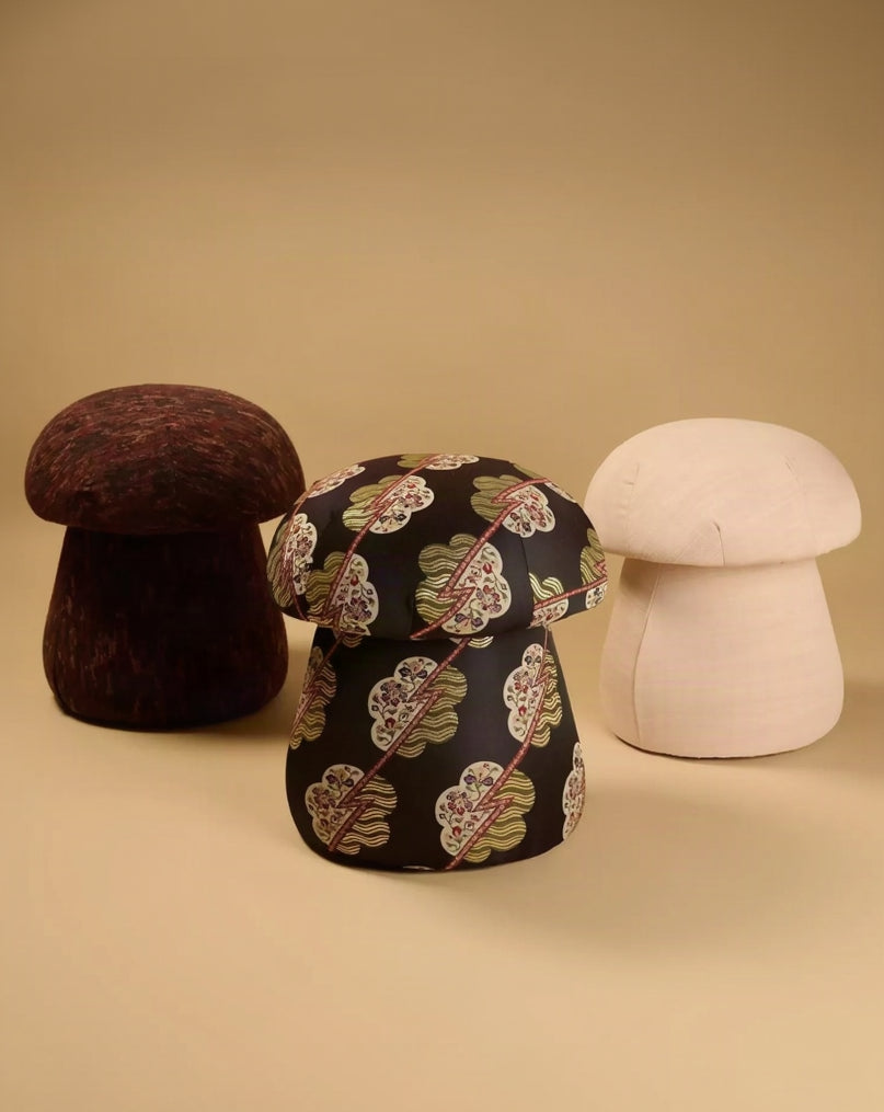 Three mushroom-shaped poufs or stools in different materials and colors: a dark brown woven one on the left, a black fabric one with a floral pattern in the center, and a smooth off-white one on the right, set against a solid beige background.