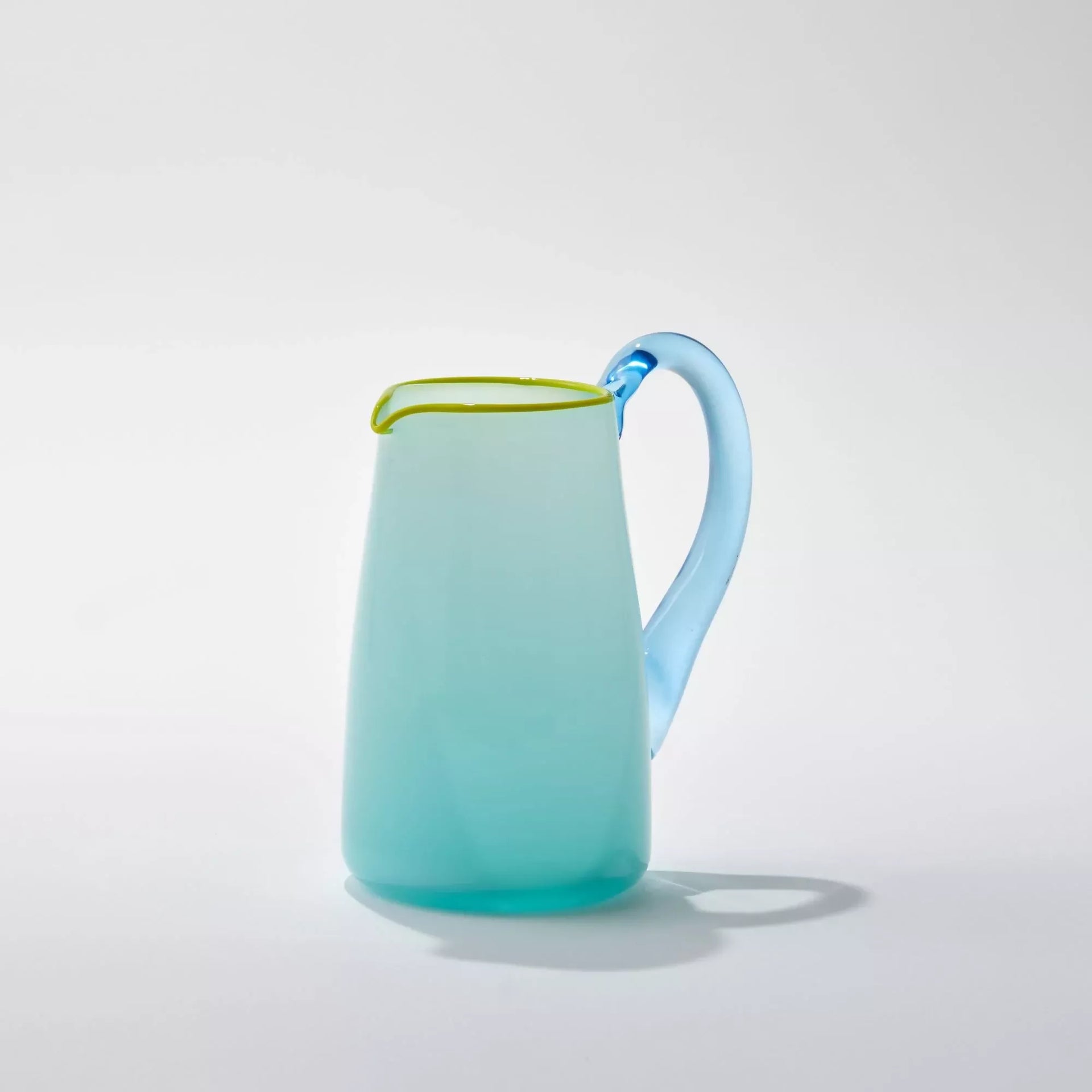 An image of the hand-blown blue glass milk jug with an aqua blue handle and a celadon lip, against a white background- available  to buy on the-salon.com. SALON curates the finest , unique, handmade pieces from the interior industry's best makers and brands. Shop luxury furniture, lighting, rugs, art, antiques and vintage pieces and accessories from SALON interior designed rooms.