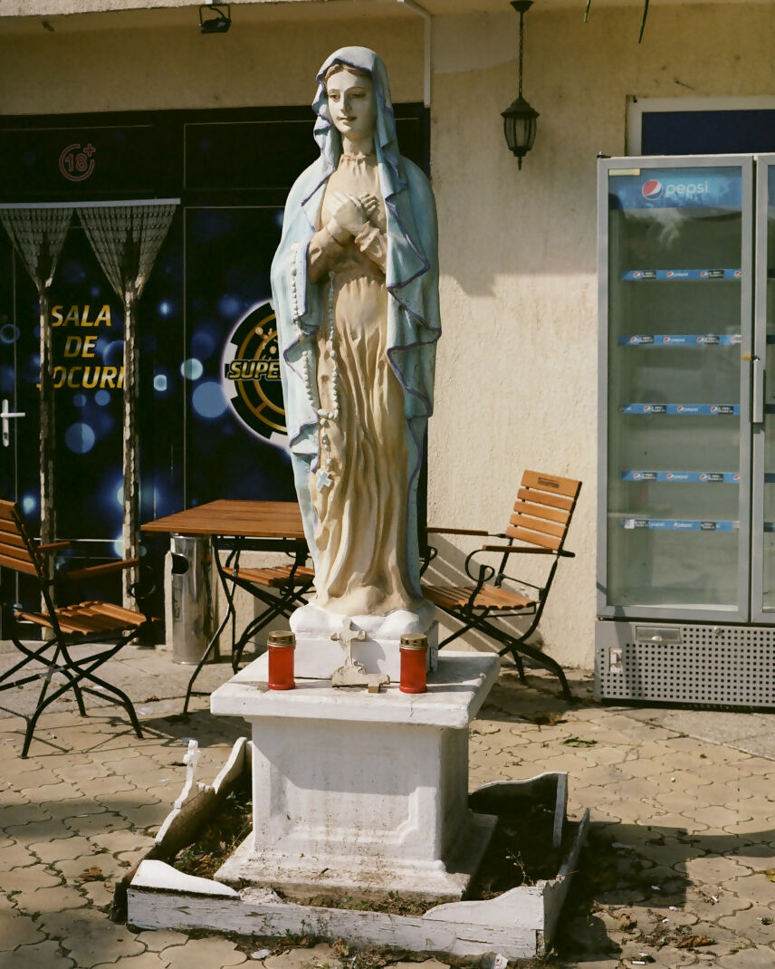 A photograph of a statue of the Virgin Mary with a blue and gold robe, standing on a white pedestal, with outdoor seating and a refrigerator in the background,  available  to buy on the-salon.com. SALON curates the finest , unique, handmade pieces from the interior industry's best makers and brands. Shop luxury furniture, lighting, rugs, art, antiques and vintage pieces and accessories from SALON interior designed rooms.