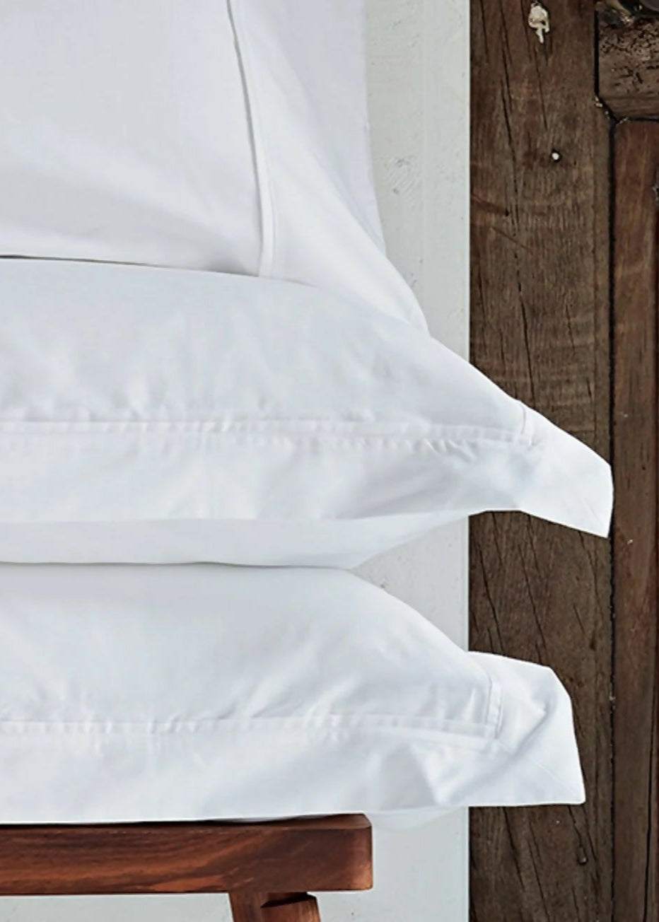 A pair of folded white organic cotton pillowcases placed on a wooden surface,  available  to buy on the-salon.com. SALON curates the finest , unique, handmade pieces from the interior industry's best makers and brands. Shop luxury furniture, lighting, rugs, art, antiques and vintage pieces and accessories from SALON interior designed rooms.