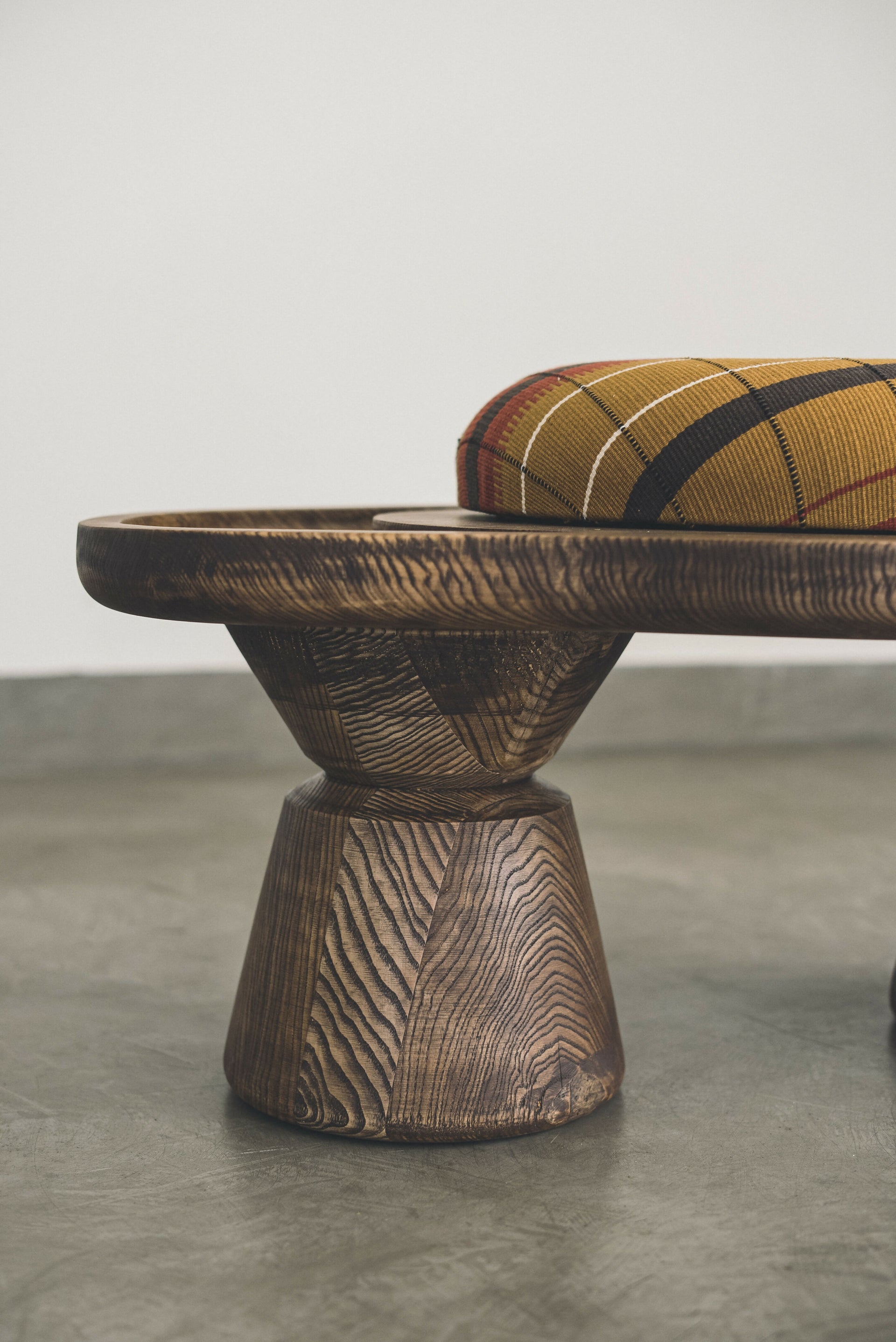 Close-up to the wood of a sculptural single-seater ottoman with a curved body, upholstered in mustard color, on a concrete floor,  available  to buy on the-salon.com. SALON curates the finest , unique, handmade pieces from the interior industry's best makers and brands. Shop luxury furniture, lighting, rugs, art, antiques and vintage pieces and accessories from SALON interior designed rooms.