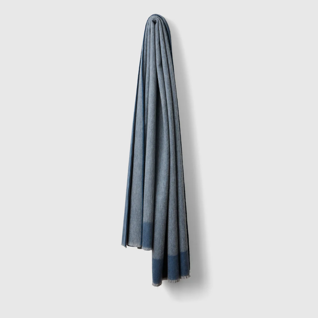 A folded slate blue cashmere throw blanket with a decorative border detail,  available  to buy on the-salon.com. SALON curates the finest , unique, handmade pieces from the interior industry's best makers and brands. Shop luxury furniture, lighting, rugs, art, antiques and vintage pieces and accessories from SALON interior designed rooms.