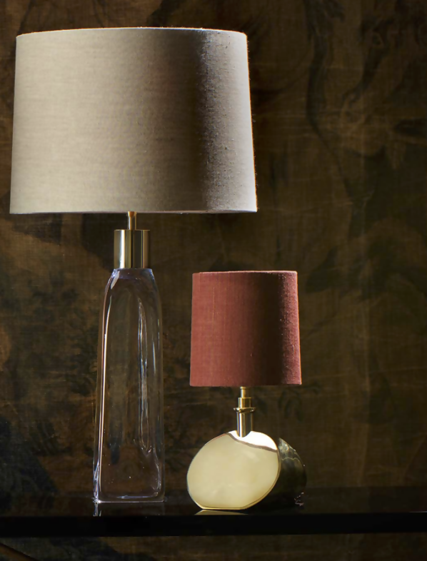 Two table lamps, one with a cinnamon brown shade and cream card lining, and the other with a clear shade, both with a gold base, against a dark background, available  to buy on the-salon.com. SALON curates the finest , unique, handmade pieces from the interior industry's best makers and brands. Shop luxury furniture, lighting, rugs, art, antiques and vintage pieces and accessories from SALON interior designed rooms.