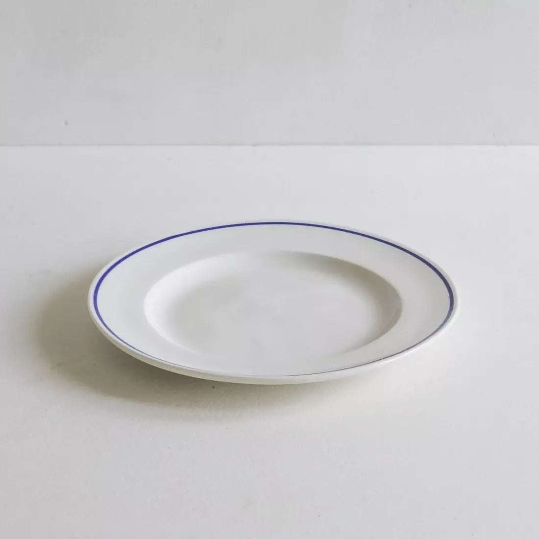 A round white porcelain side plate with a rich cobalt blue line along the rim, displayed against a white background, available  to buy on the-salon.com. SALON curates the finest , unique, handmade pieces from the interior industry's best makers and brands. Shop luxury furniture, lighting, rugs, art, antiques and vintage pieces and accessories from SALON interior designed rooms