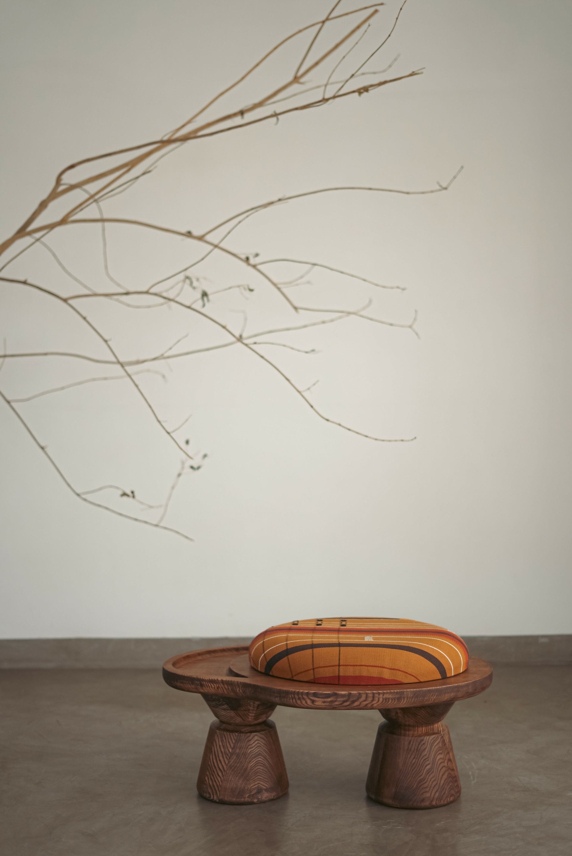 A sculptural single-seater ottoman with a curved body, upholstered in mustard color, placed next to a large ceramic pot with a branch in it, on a concrete floor,  available  to buy on the-salon.com. SALON curates the finest , unique, handmade pieces from the interior industry's best makers and brands. Shop luxury furniture, lighting, rugs, art, antiques and vintage pieces and accessories from SALON interior designed rooms.