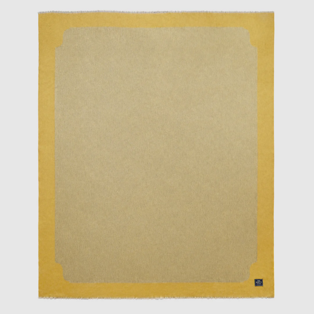 A folded gold-colored cashmere throw blanket with a decorative border, displaying a small blue and silver brand tag on the bottom right corner,  available  to buy on the-salon.com. SALON curates the finest , unique, handmade pieces from the interior industry's best makers and brands. Shop luxury furniture, lighting, rugs, art, antiques and vintage pieces and accessories from SALON interior designed rooms.